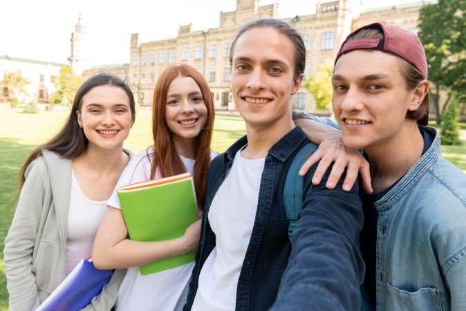 Θεματική εικόνα για το τι πρέπει να ξέρω για το Erasmus. Τέσσερις χαμογελαστοί φοιτητές βγάζουν selfie στο πανεπιστήμιό τους.
