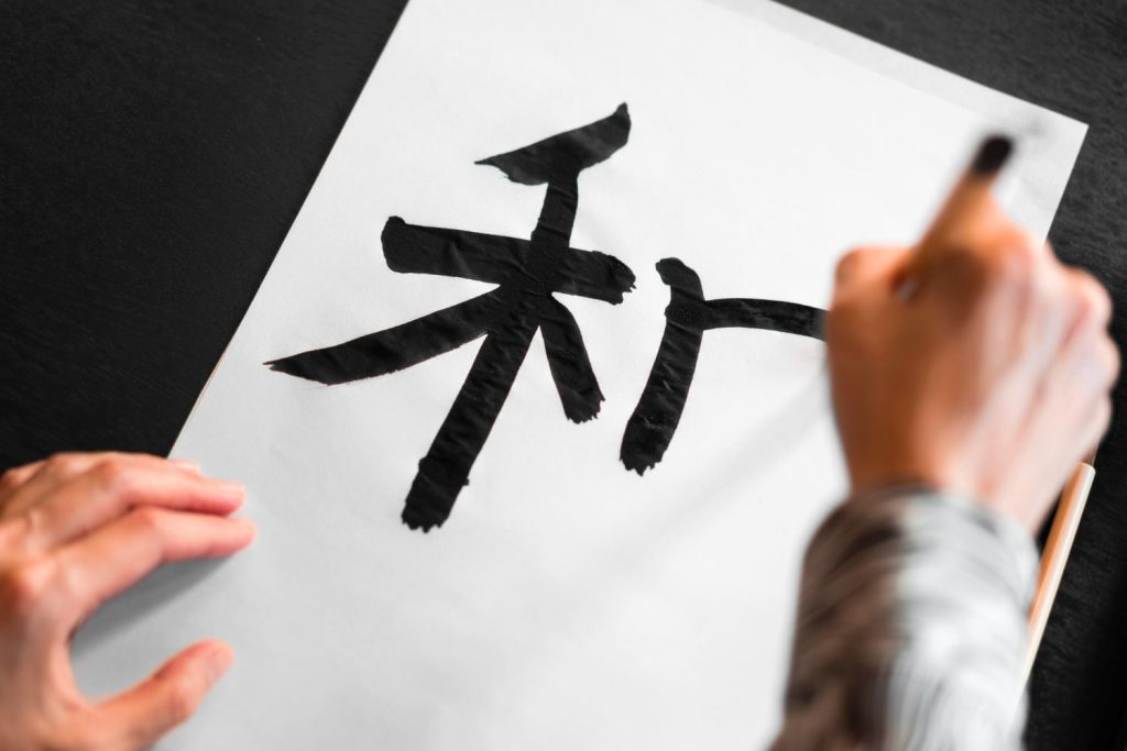 Άτομο σχηματίζει με πινέλο κινεζικά ιδεογράμματα μαύρου χρώματος, πάνω σε μεγάλο λευκό χαρτί.